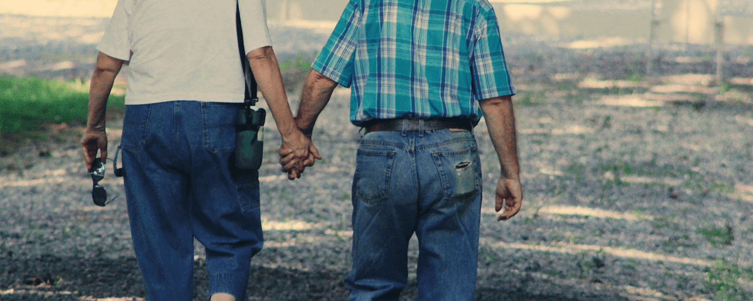 An elderly couple walks hand in hand down a gravel path. The woman wears a white shirt, hat, and blue scarf, while the man wears a plaid shirt and cap. A wooden fence is visible in the background.